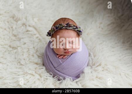 Schlafendes, zehntägiges, neugeborenes Mädchen, das in einer lilafarbenen Umhüllung schwamm. Im Studio auf einem weißen Schaffellrug gedreht Stockfoto