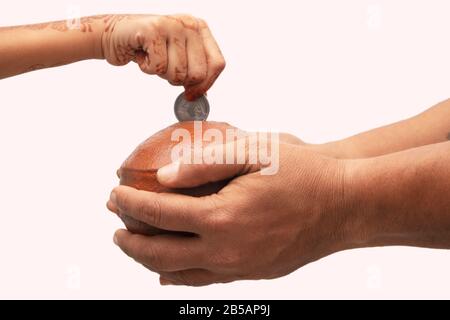 Hände, die indische Penny Bank mit Lehm und Kinderhand halten, die auf Isoliertem Hintergrund eine Münze in den Erdtopf oder die Piggy-Bank aus Terrakotta hinzufügt - Stockfoto