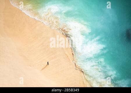 Blick von oben, atemberaubender Blick auf eine Person, die bei Sonnenuntergang an einem schönen Strand spaziert, der von einem türkisfarbenen Meer umspült wird. Kelingking Beach, Nusa Penida. Stockfoto