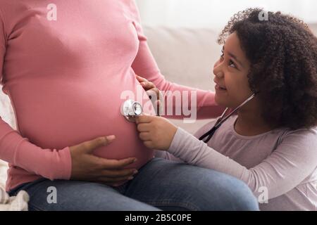 Kleine afro-Tochter, die mit Stethoskop dem Bauch ihrer schwangeren Mutter lauscht Stockfoto