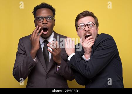 In einer schwarzen Suite, die an der gelben Wand isoliert ist, haben afrikanische und kaukasische Geschäftsleute Angst. Finanzkrisenkonzept. Stockfoto