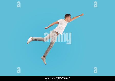 Durchgehendes Porträt eines motivierten selbstbewussten Mannes in weißem T-Shirt und lässigen Hosen, die in der Luft auffliegen, als Superheld mit erhobener Hand, der Übermacht spürt. Stockfoto