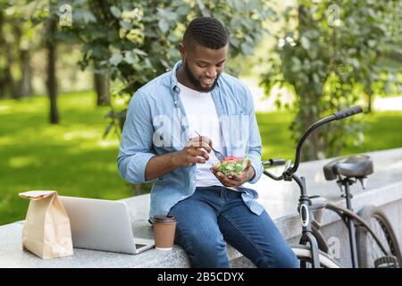 Schwarzer Geschäftsmann, der Salat zum Mittagessen hat und im Freien auf einem Laptop arbeitet Stockfoto