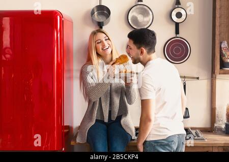 Spaß beim Frühstück. Junge Frau, die ihren Mann mit Croissant in der Küche füttert Stockfoto