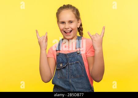 Portrait des überfröhlichen kleinen Mädchens mit Geflecht in Denim-Overalls, das Rock- und Roll-Geste macht und Yeah schreit, es ist verrückt, mit h cooles Rock-Zeichen zu zeigen Stockfoto