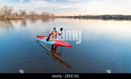 Der ältere männliche Paddler in einem Trockenanzug und einer Life-Jacke beendet sein Training mit einem langen Rennstativ auf einem Paddleboard an einem See in Colorado, im Winter oder im Ohr Stockfoto