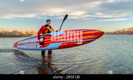 Der ältere männliche Paddler in einem Trockenanzug und einer Life-Jacke beendet sein Training mit einem langen Rennstativ auf einem Paddleboard an einem See in Colorado, im Winter oder im Ohr Stockfoto