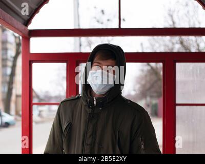 Kerl in einer medizinischen Maske steht an einer Bushaltestelle. Isoliert, um Viruserkrankung zu vermeiden, Kovid 19 Masern Grippe Windpocken kalt. Stockfoto