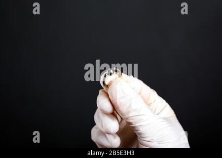 Hand in Weiß hält den durchstechenden Ohrring in silberner Farbe auf dunklem Hintergrund in Nahaufnahme. Ohrschmuck. Stockfoto