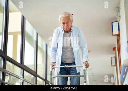 asiatischer Mann, der mit einem Geher im Flur des Krankenhauses oder Pflegeheims spazieren ging Stockfoto