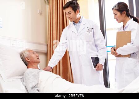 Älterer asiatischer Mann, der auf dem Bett liegt und mit Ärzten in der Krankenstation spricht Stockfoto