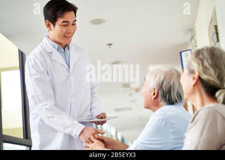 Freundlicher junger asiatischer Arzt, der im Wartebereich mit älteren Patienten im Krankenhaus die Hände schüttelt Stockfoto