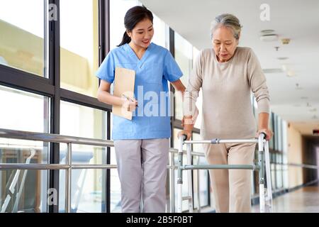 Junger asiatischer Physiotherapeut, der mit einer älteren Frau beim gehen mit einem Wanderer arbeitet Stockfoto