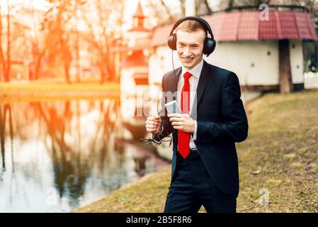 Lustiger junger Kerl hört Musik mit großen Kopfhörern. Ein Mann in schwarzem Anzug und roter Krawatte. Geschäftsmann ruht, entspannt. Getönt Stockfoto