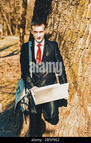 Porträt eines Studenten in schwarzem Anzug und roter Krawatte an der Natur in der Nähe eines großen Baumes. Auf der Schulter einer jungen Kerltasche. Freiberufler hält einen Laptop Stockfoto