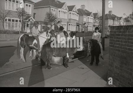 1970er Jahre, historisch, in einer Vorstadtstraße, halten Jugendliche auf Pferden und Ponys an einem Wassertrog, um ihren Tieren Erfrischung zu geben, South London, England, während eines besonders heißen Sommers. Im Laufe des Jahrzehnts waren einige von ihnen extrem heiß. Stockfoto