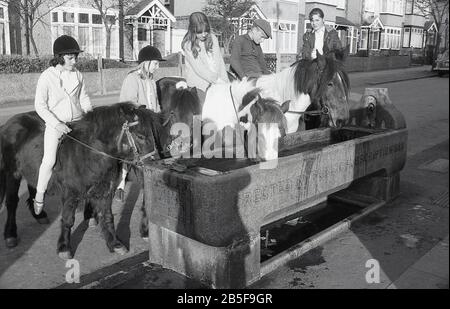 In den 1970er Jahren, historisch, halten Jugendliche auf Pferden und Ponys an einem alten Wassertrog aus Stein - erbaut im Jahr 1888 -, um ihren Tieren während eines heißen Sommers Erfrischung zu geben, South London, England. Im Laufe des Jahrzehnts waren einige der Sommer extrem heiß. Stockfoto