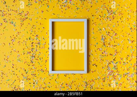 Weißer Rahmen und bunte Einstreuungen für Kuchen auf gelbem Hintergrund. Geburtstagsparty-Konzept. Verspotten Sie sich, platzieren Sie Text. Stockfoto