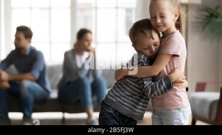 Gestresstes kleines Mädchen, das kleineren Bruder umarmt und die Eltern überwunden hat, argumentiert die Scheidung. Stockfoto