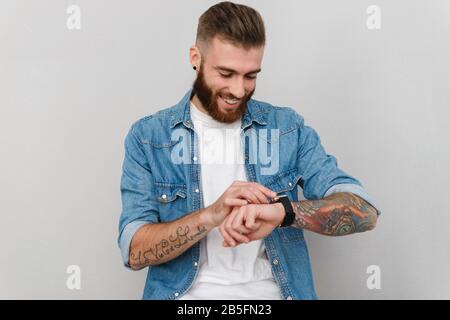 Porträt eines gutaussehenden jungen Mannes mit legerer Kleidung, die isoliert über grauem Hintergrund steht und die Zeit auf seiner Armbanduhr überprüft Stockfoto