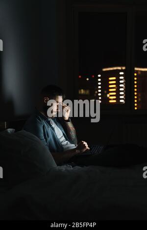 Müde junge bärtige Mann, der nachts zu Hause auf dem Bett liegt und am Laptop arbeitet Stockfoto