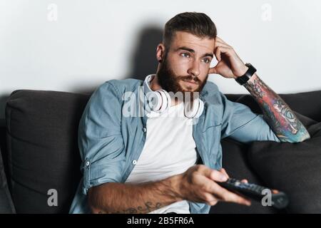 Attraktiver gelangweilter junger bärtiger Mann, der sich zu Hause auf einer Couch entspannt und Fernsehen kann Stockfoto