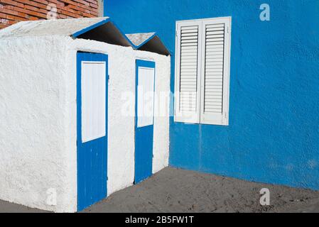 Badekabinen in Beton, pastellblau und weiß lackiert neben blauer Wand mit weißem Fenster mit geschlossenen Fensterläden Stockfoto