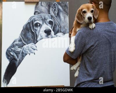 Ein Acrylgemälde, das ein Mann mit einem Beagle-Hund auf weißer Leinwand trägt, während ein Künstler einen Hund an der Vorderseite seiner Arbeit trägt. Stockfoto