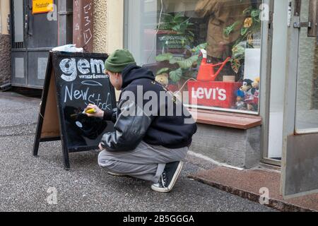 Junger Mann, der auf einer Tafel auf dem Bürgersteig vor Vintage und 2. Hand Shop Gem im finnischen Kallio Viertel in Helsinki schreibt Stockfoto