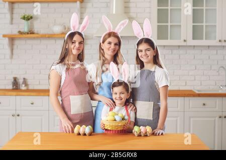 Frohe ostern. Mutter und Töchter mit Kaninchenohren schmücken Ostereier, die an einem Tisch in der Küche sitzen. Stockfoto