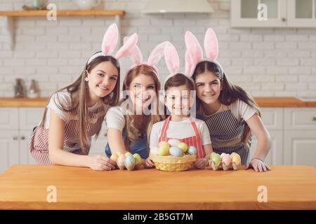 Frohe ostern. Mutter und Töchter mit Kaninchenohren schmücken Ostereier, die an einem Tisch in der Küche sitzen. Stockfoto