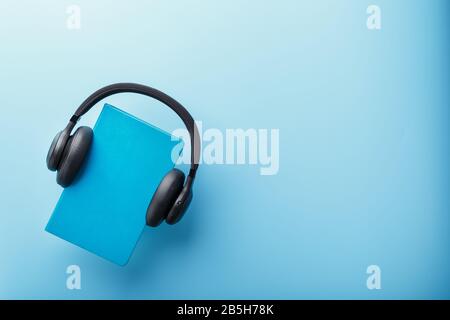 Kopfhörer werden an einem Buch in blauer Hardcover auf blauem Hintergrund und in der Draufsicht getragen. Das Konzept von Hörbuch und Fernunterricht, E-Learning. SP kopieren Stockfoto