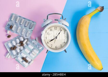 Medizinische Pillen in Blisterpackungen und Bananen mit einem weißen klassischen Wecker auf einem rosa und hellblauen kreativen Hintergrund. Das Konzept der geschmeidigen Ernährung Stockfoto