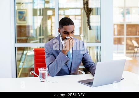 Porträt eines kranken jungen Mannes im Business-Anzug, der eine Kälte hatte und seine Nase wehte, während er hinter dem Schreibtisch bei der Arbeit mit dem Computer saß Stockfoto