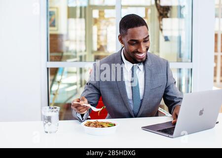 Porträt eines fröhlichen lächelnden jungen Mannes in Business-Anzug Mittagessen an seinem Schreibtisch essen, während er an einem Laptop in seinem Büro arbeitet Stockfoto