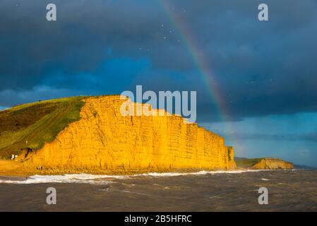 West Bay, Dorset, Großbritannien. März 2020. Wetter in Großbritannien. Ein Regenbogenbogen über den Klippen, der am späten Nachmittag bei Sonnenschein in der West Bay in Dorset als dunkle Duschwolke beleuchtet wird, geht über. Bildnachweis: Graham Hunt/Alamy Live News Stockfoto
