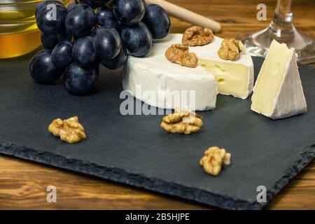 Camembert Käse in einem dunklen Tablett neben süßen blauen Trauben Nüsse und Honig auf Holzgrund Stockfoto