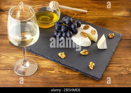 Camembert-Käse, Nüsse, Honig und süße Trauben auf dem Hintergrund eines Glases aus weißem Trockenwein und Holzhintergrund mit Copyspace Stockfoto