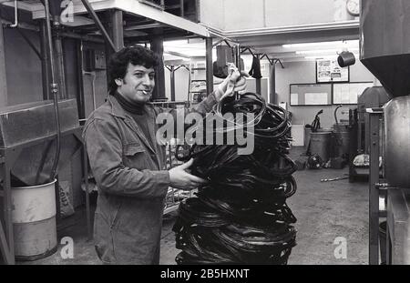 In den 1970er Jahren bleibt ein historischer, männlicher Arbeiter mit Zigarette im Mund und in Overalls mit einem Turm aus Kunststoff oder PVC-Abfall nach dem Pressen von Schallplatten oder LPs, USA, zurück. Die Produktion von Schallplatten aus Vinyl war eine unordentliche, schmutzige Angelegenheit, bei der große Mengen von Kunststoffpolymer aus raffiniertem Öl beteiligt waren. Wenn dieses Restmaterial auf Deponien entsorgt würde, würde es bis zu 1.000 Jahre dauern, bis es zersetzt ist. Stockfoto