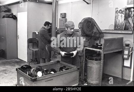 1970er Jahre, historischer, männlicher Arbeiter in Overalls mit Plastikmüll, Überreste von weggeworfenen Schallplatten, USA. Die Produktion von Vinylplatten war eine schmutzige Angelegenheit, bei der es um ölbasierten Kunststoff ging. Stockfoto