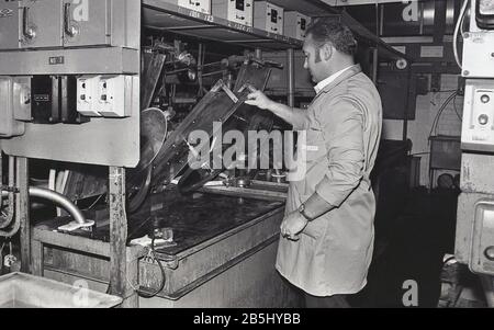 1970er Jahre, historisch, Plattenherstellung, ein männlicher Arbeiter in einem Gesamt, der Maschinen verwendet, um die Kunststoff-Vinyl, die bei der Herstellung oder Pressung von Vinyl-Platten verwendet werden, USA. Stockfoto