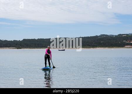 Foz Do Arelho, Portugal, 8. März 2020 - ältere Frau auf einem SUP-Paddlebarden. Mit schwarzem und pinkfarbenem Neoprenanzug Stockfoto