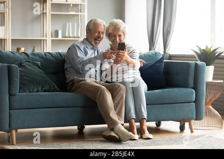 Aufgeregte 60er-Jahre-Paare sitzen auf der Couch und machen Selfies auf dem Handy Stockfoto