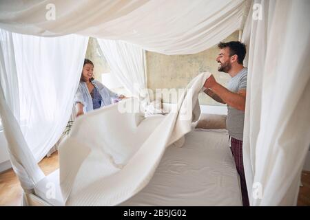 Das junge Paar spreizt mit Freude Bettlaken im Schlafzimmer: Schlafzimmer, Morgen, Routine, Zusammengehörigkeit, Glückskonzept Stockfoto