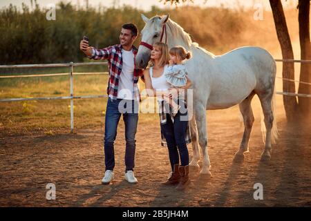 Lächelnde junge Familie, die auf dem Land auf der Pferdefarm genießt und selfie macht. Stockfoto