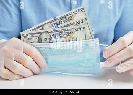 Eine Hand in Handschuhen hält eine schützende medizinische Maske mit Dollar. Das Konzept des Unternehmens und viel Geld für den Schutz. Nahaufnahme. Stockfoto