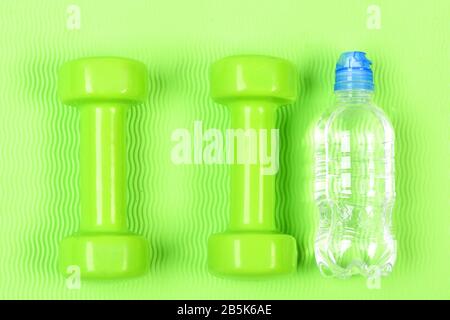 Sport-, Energie- und Ernährungskonzept. Zwei grüne Fitness-Hanteln und eine Flasche Wasser auf grünem Hintergrund. Zwei Kunststoff-Hanteln und eine durchsichtige Flasche. Handgewichte und Wasser auf hellem Hintergrund. Stockfoto
