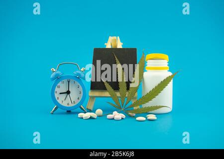 Medizinisches Marihuana-Konzept mit frischen grünen Blättern, pharmazeutischen Flaschen und Pillen, Wecker und kleiner Kreideplatte über blauem Hintergrund mit Kopie Stockfoto