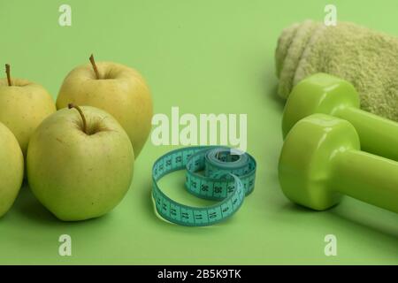 Gesunde Regimegeräte. Barglocken aus Kunststoff in der Nähe saftiger grüner Äpfel. Hanteln in hellgrüner Farbe, Cyan messen Band, Handtuch und Obst auf grünem Hintergrund. Leichtathletik und Gewichtsverlust Konzept Stockfoto