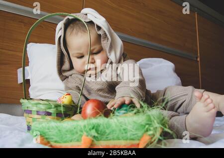 Süßer Babyjunge im Hunny Kostüm, der mit dem Korb mit bunten ostereiern spielt. Ostereierjagd und Babyentwicklung. Stockfoto